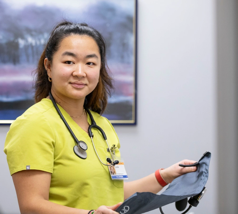 A healthcare professional with shoulder-length hair, wearing a yellow scrub top, a stethoscope around her neck, and a name badge reading "Julia". She is holding a blood pressure cuff and is standing in front of a wall with a framed landscape painting, suggesting she is in a medical office or clinic setting.