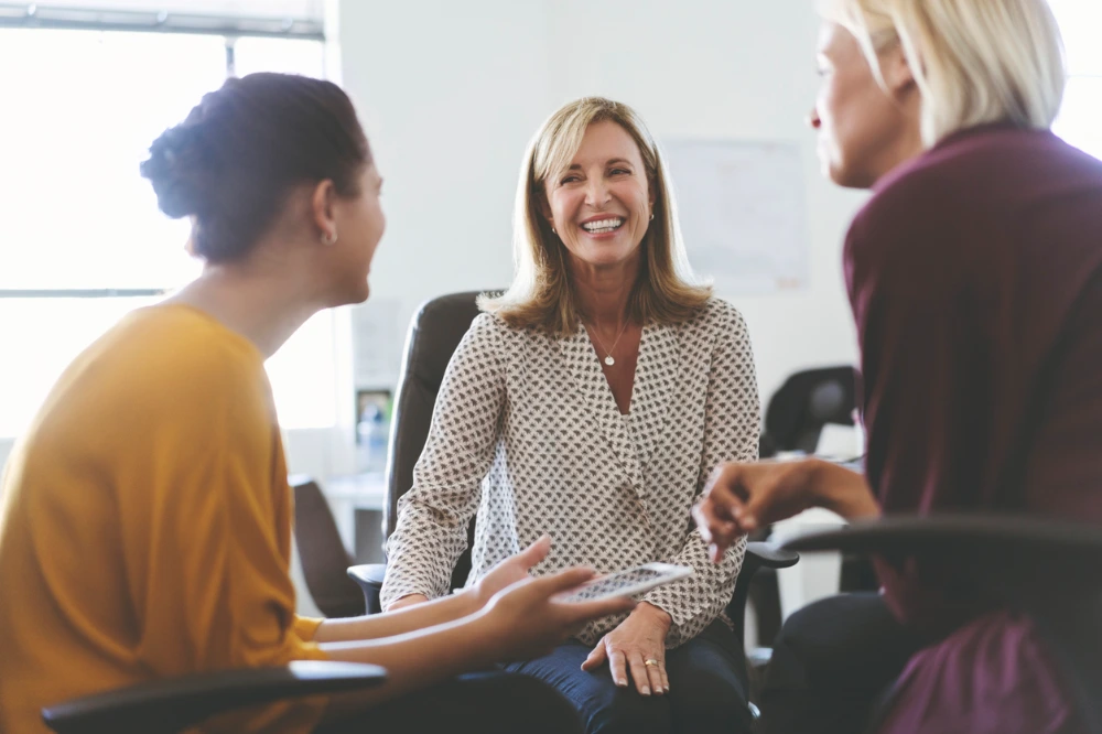 A group of three professionals in a meeting, with two women engaged in conversation while a third person listens.