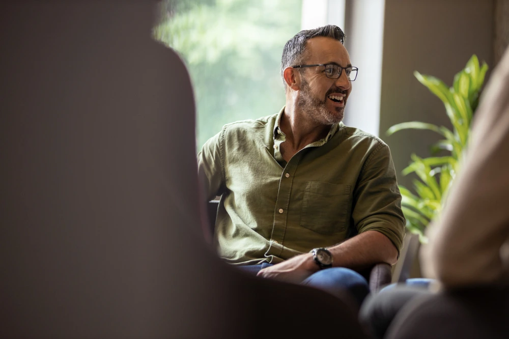 A man in a green shirt laughing and talking in a bright room with soft focus on foreground and clear view of his face.