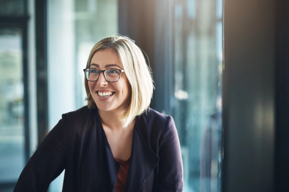 A cheerful woman with glasses smiling broadly, seated indoors with soft lighting and a blurred background.