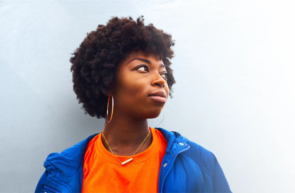 A young woman with a thoughtful expression stands against a light-colored wall. She has natural curly hair and wears hoop earrings, a bright orange shirt, and a blue jacket. She is looking off to the side, creating a sense of contemplation or daydreaming.