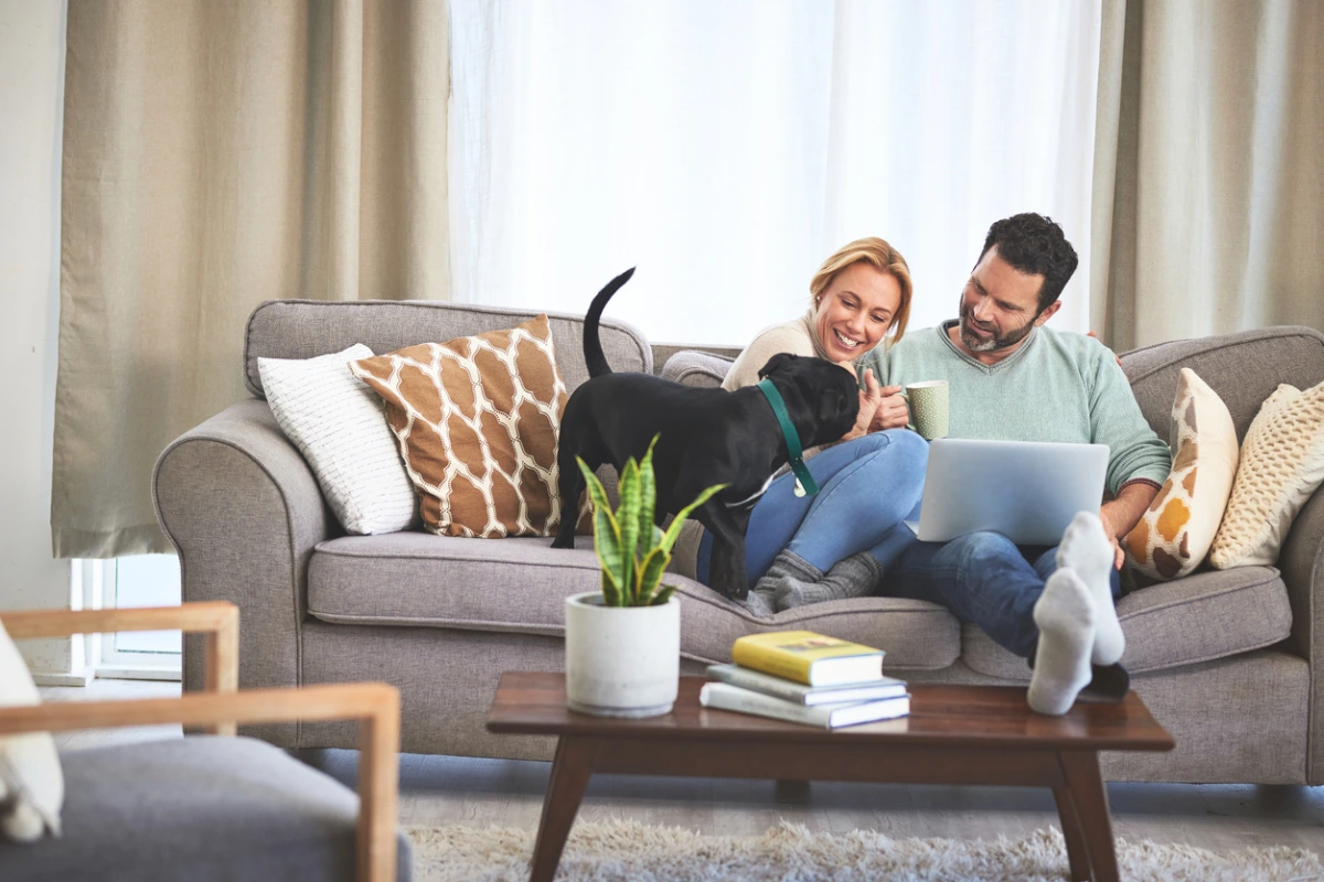 A couple sitting on a couch with a dog, enjoying coffee and using a laptop, in a cozy living room setting.