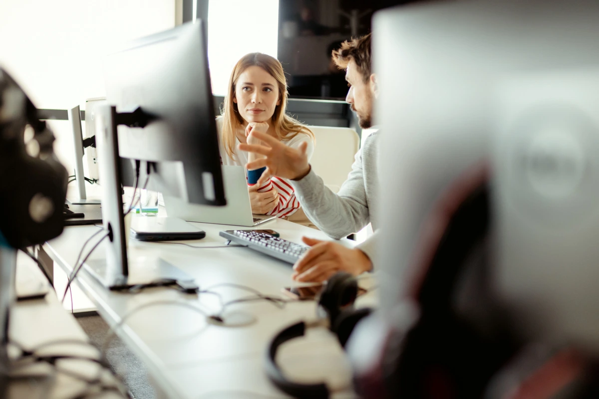 Two coworkers discussing work at a desk with multiple computer monitors. A blonde woman listens attentively while a man gestures, explaining something. The office setting is modern with technology visible.