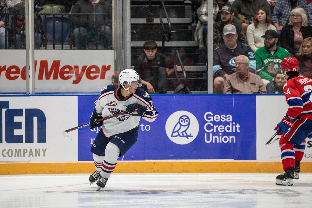 Hockey player in white jersey skating on ice during a game, with spectators and advertisements visible in the background