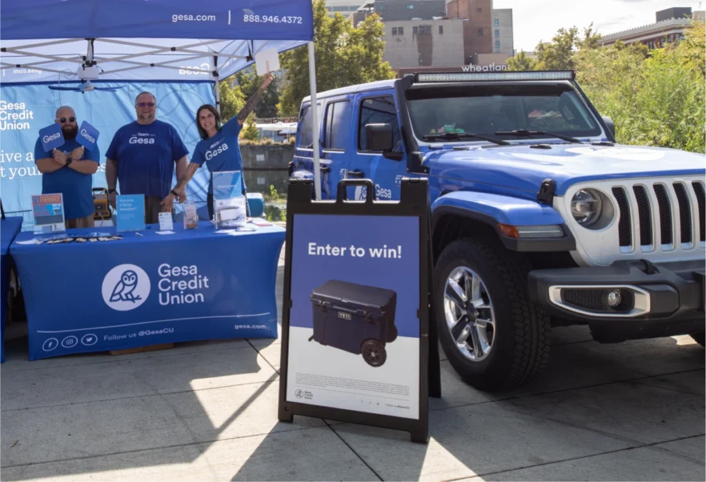 Image of a Gesa Credit Union promotional booth set up outdoors, with a blue Jeep parked next to it. The booth has a blue tent with the Gesa logo, and three smiling people in blue Gesa t-shirts are standing behind a table with promotional materials. A sign on the booth reads "Enter to win!" next to an image of a Yeti cooler, the prize for the giveaway.