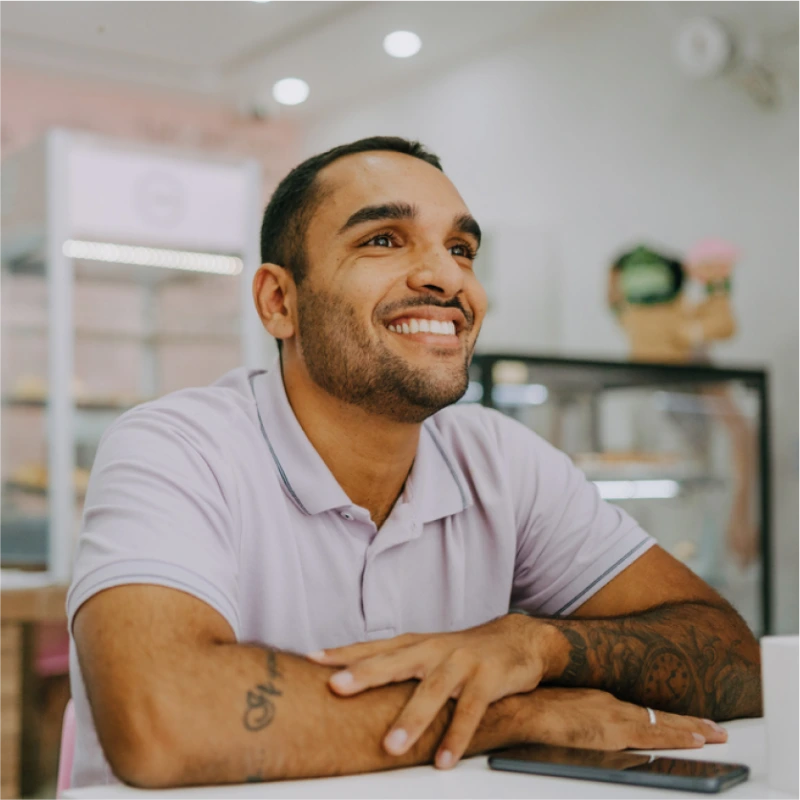 A smiling man with short dark hair and tattoos on his forearm sits at a table in a bright cafe, wearing a light purple polo shirt. Behind him, there are refrigerated display cases filled with pastries and cakes.